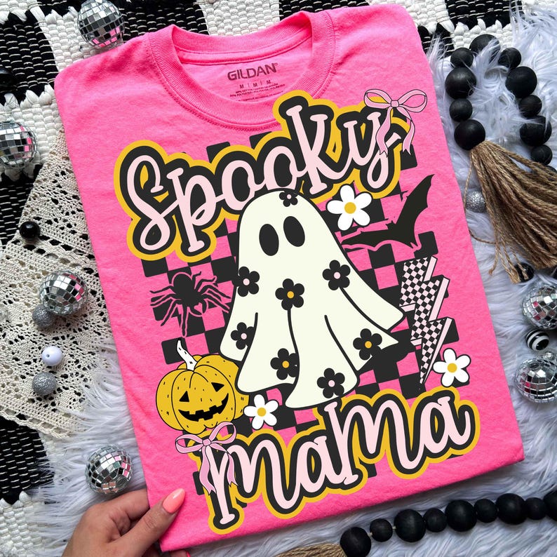 Puede incluir: Camiseta rosa brillante con un dise&ntilde;o de Halloween. El dise&ntilde;o presenta las palabras "Spooky Mama", un fantasma, una calabaza, una ara&ntilde;a y un murci&eacute;lago. La camiseta tiene cuello redondo y mangas cortas.