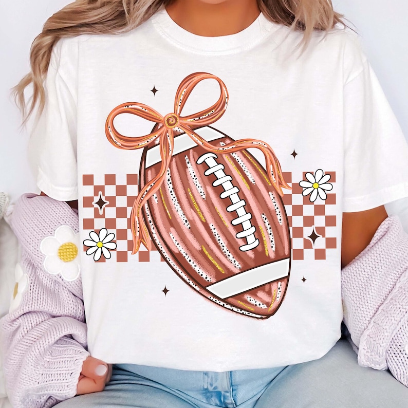 May include: White t-shirt featuring a football graphic with a brown bow, white laces, and gold accents. The design includes a brown and white checkered pattern, daisies, and starbursts. A casual, sporty design.