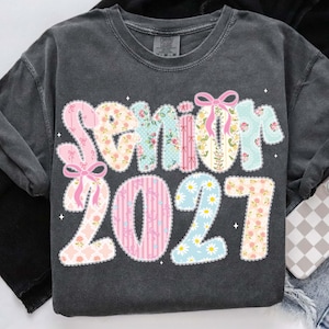 May include: Dark gray sweatshirt with the word "Senior" and the number "2021" in a floral and patterned design. The letters and numbers are in pastel colors, with pink bows and daisy accents.