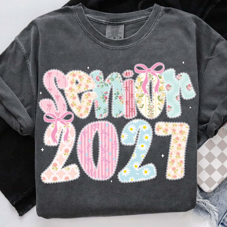 May include: Dark gray sweatshirt with the word "Senior" and the number "2021" in a floral and patterned design. The letters and numbers are in pastel colors, with pink bows and daisy accents.