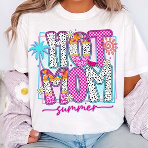 Puede incluir: Camiseta blanca con un gr&aacute;fico colorido que dice "HOT MOM summer". El dise&ntilde;o incluye una palmera, flores, un sol y una rodaja de sand&iacute;a. Las letras est&aacute;n en varios patrones y colores.