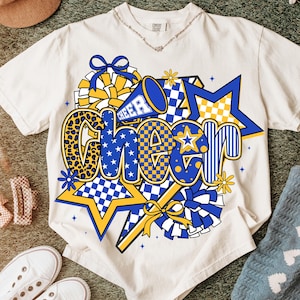May include: Cream-colored t-shirt with a vibrant "CHEER" graphic. The design features blue and yellow stars, checkered patterns, and a megaphone. The word "CHEER" is in a mix of fonts and patterns, including leopard print.