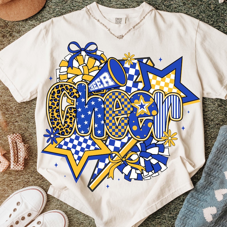 May include: Cream-colored t-shirt with a vibrant "CHEER" graphic. The design features blue and yellow stars, checkered patterns, and a megaphone. The word "CHEER" is in a mix of fonts and patterns, including leopard print.