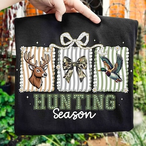 Puede incluir: Sudadera negra con un ciervo, un lazo de camuflaje y una ilustración de pato. Las palabras "HUNTING Season" están impresas en verde y blanco. El diseño está enmarcado con patrones florales y a rayas.