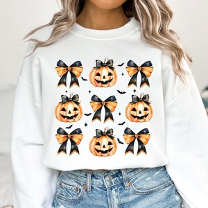 May include: White sweatshirt with a pattern of orange and black jack-o'-lantern pumpkins with black bows and gold glitter accents.