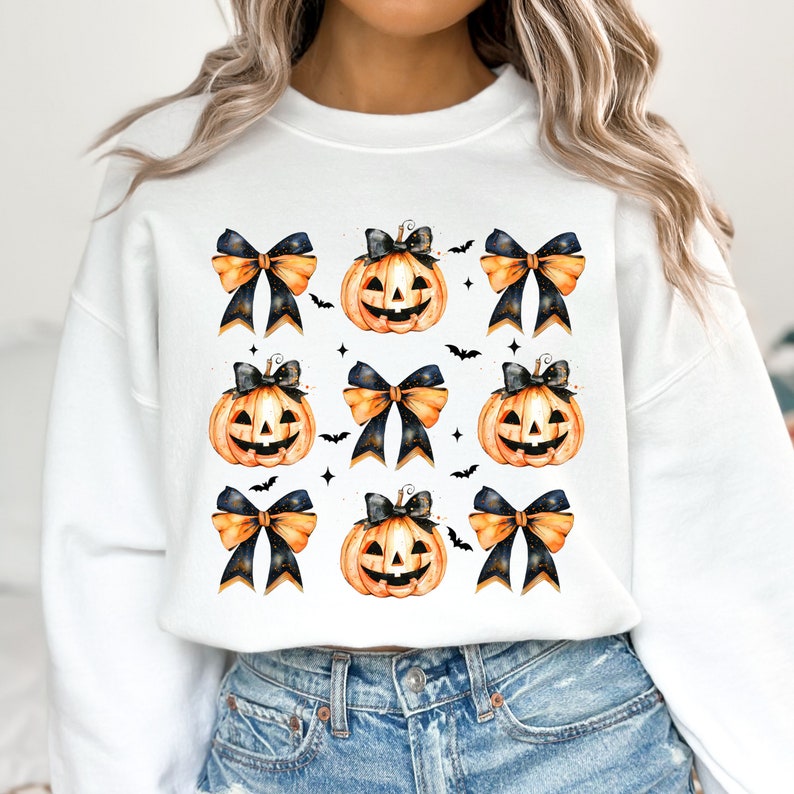 May include: White sweatshirt with a pattern of orange and black jack-o'-lantern pumpkins with black bows and gold glitter accents.