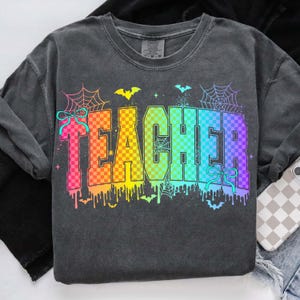 May include: Dark gray sweatshirt with the word "TEACHER" in a rainbow checkered design. The design includes bats, spiderwebs, and dripping paint, creating a Halloween theme.