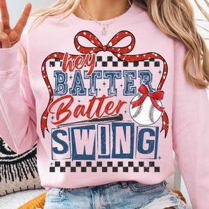 May include: Pink sweatshirt with a red and white bow, baseball bat, and baseball. The text on the sweatshirt says "Hey Batter Batter Swing".