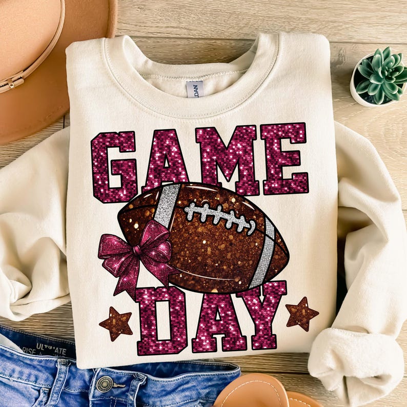 Puede incluir: Sudadera de color crema con las palabras "GAME DAY" en letras rosas brillantes. Un bal&oacute;n de f&uacute;tbol con un lazo rosa est&aacute; en el centro. Dos estrellas rosas est&aacute;n en la parte inferior. La sudadera est&aacute; doblada sobre una superficie de madera.
