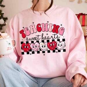 Pu&ograve; includere: Felpa rosa con il testo "TEACHING sweet hearts" in un font retr&ograve;. Cinque cuori di cartoni animati con diversi motivi sono allineati sotto il testo.