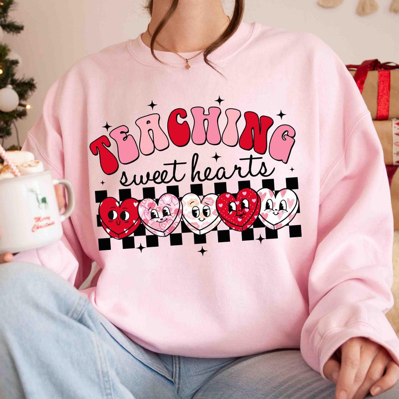 Pu&ograve; includere: Felpa rosa con il testo "TEACHING sweet hearts" in un font retr&ograve;. Cinque cuori di cartoni animati con diversi motivi sono allineati sotto il testo.