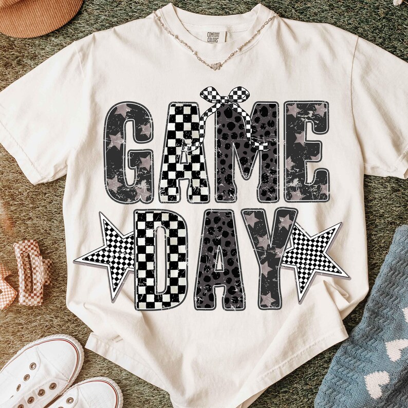 May include: Cream-colored t-shirt with the words "GAME DAY" in a distressed, black and gray design. The letters have a checkered pattern and star accents. A checkered bow sits above the word "GAME".