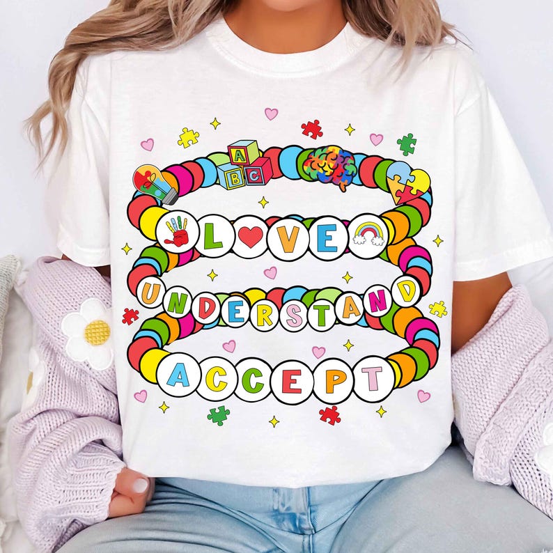 May include: White t-shirt with a colorful design featuring the words "LOVE UNDERSTAND ACCEPT" in a circle of beads. The design also includes a rainbow, a puzzle piece, a lightbulb, and a block with the letters "ABC".
