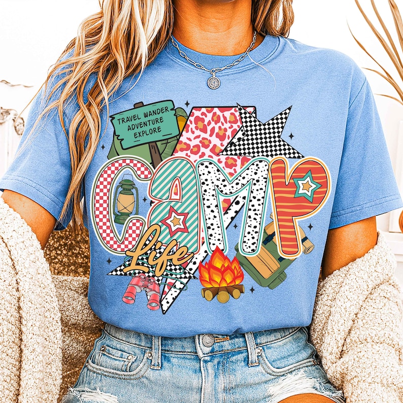 Puede incluir: Una camiseta azul claro con un dise&ntilde;o gr&aacute;fico colorido que dice "Camp Life". El gr&aacute;fico incluye una tienda de campa&ntilde;a, una linterna, una fogata, binoculares y una mochila. El texto "Travel Wander Adventure Explore" est&aacute; en un cartel dentro del gr&aacute;fico.