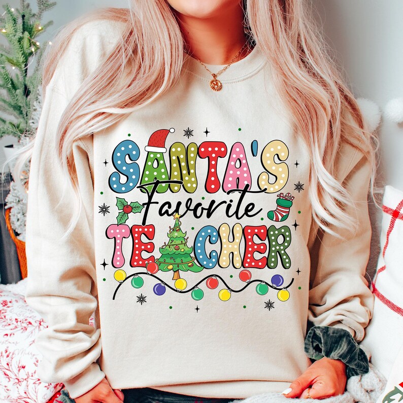 May include: A cream-colored sweatshirt with a festive Christmas design. The text "SANTA'S FAVORITE TEACHER" is written in colorful, polka-dotted letters with a Christmas tree and other holiday decorations.