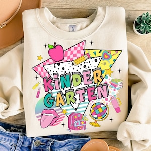 K&ouml;nnte beinhalten: Ein beiger Pullover mit einem farbenfrohen Grafikdesign, das das Wort "KINDERGARTEN" im Retro-Stil zeigt. Das Design beinhaltet einen roten Apfel, einen Bleistift, einen Globus, einen Rucksack und andere schulthemenbezogene Elemente.