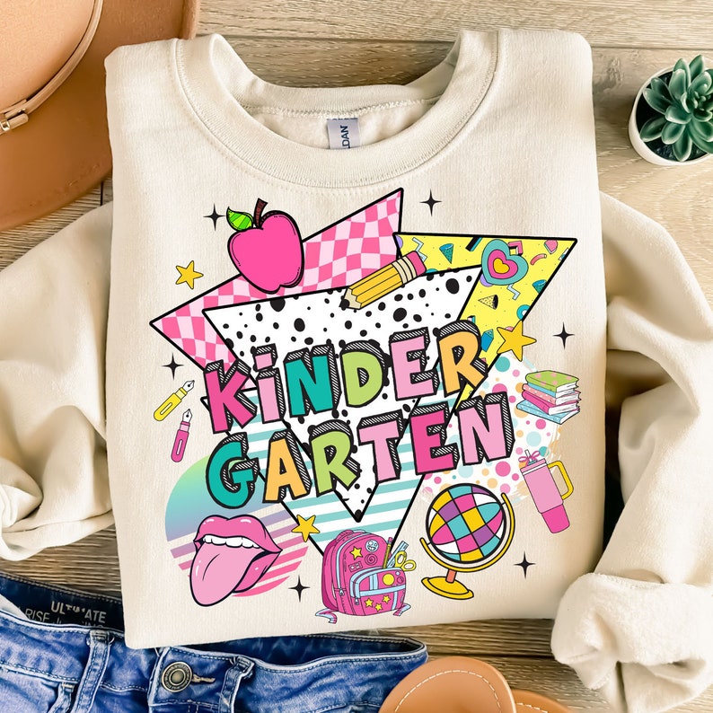 K&ouml;nnte beinhalten: Ein beiger Pullover mit einem farbenfrohen Grafikdesign, das das Wort "KINDERGARTEN" im Retro-Stil zeigt. Das Design beinhaltet einen roten Apfel, einen Bleistift, einen Globus, einen Rucksack und andere schulthemenbezogene Elemente.