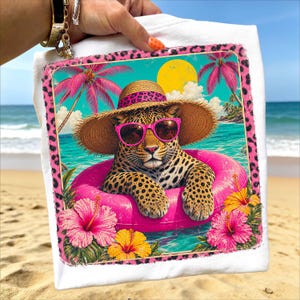 Könnte beinhalten: Weißes T-Shirt mit einem Leoparden, der eine Sonnenbrille, einen Strohhut und einen rosa Schwimmring trägt. Das Design zeigt rosa Hibiskusblüten, Palmen und einen Leopardenmuster-Rand vor einer Strandkulisse.