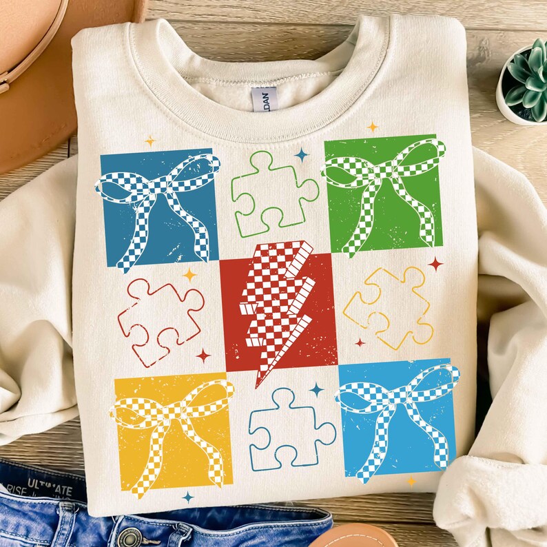 May include: A cream-colored sweatshirt with a colorful design featuring checkered bows, lightning bolts, and puzzle pieces. The design is in a patchwork style with each element in a different color.