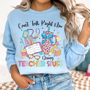 May include: Light blue sweatshirt with the text "Can't Talk Right Now Doing Teacher Stuff" in colorful letters. The design includes a coffee cup, books, pencils, and glasses.