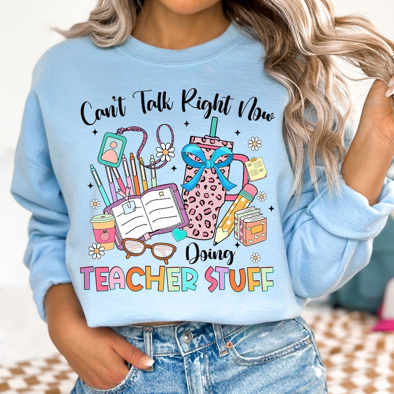 May include: Light blue sweatshirt with the text "Can't Talk Right Now Doing Teacher Stuff" in colorful letters. The design includes a coffee cup, books, pencils, and glasses.