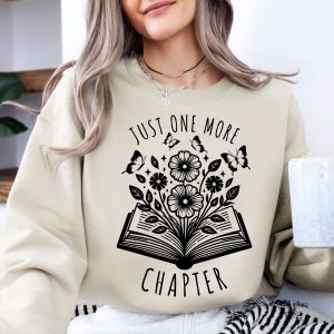 Pode incluir: Uma camisa de moletom bege com um design gráfico preto de um livro aberto com flores e borboletas. O texto "Just One More Chapter" está impresso na camisa de moletom.