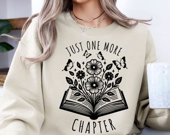Può includere: Una felpa beige con un design grafico nero di un libro aperto con fiori e farfalle. Il testo "Just One More Chapter" è stampato sulla felpa.