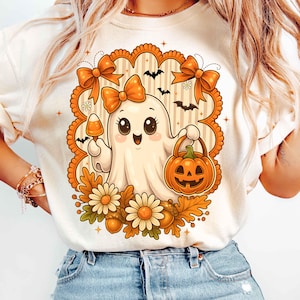 Pode incluir: Camiseta creme com um fantasma de desenho animado fofo segurando uma ab&oacute;bora e um doce de milho. O fantasma &eacute; cercado por la&ccedil;os laranja, morcegos, flores e folhas. Uma camiseta gr&aacute;fica com tema de Halloween.