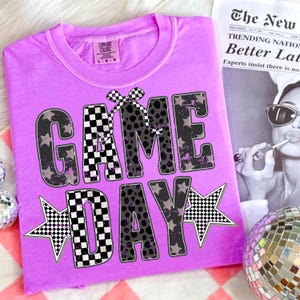 May include: A lilac Comfort Colors t-shirt with the words "GAME DAY" in a mix of black and white checkered and leopard print lettering. The design includes star accents and a checkered bow. A newspaper with the headline "The New" is in the background.