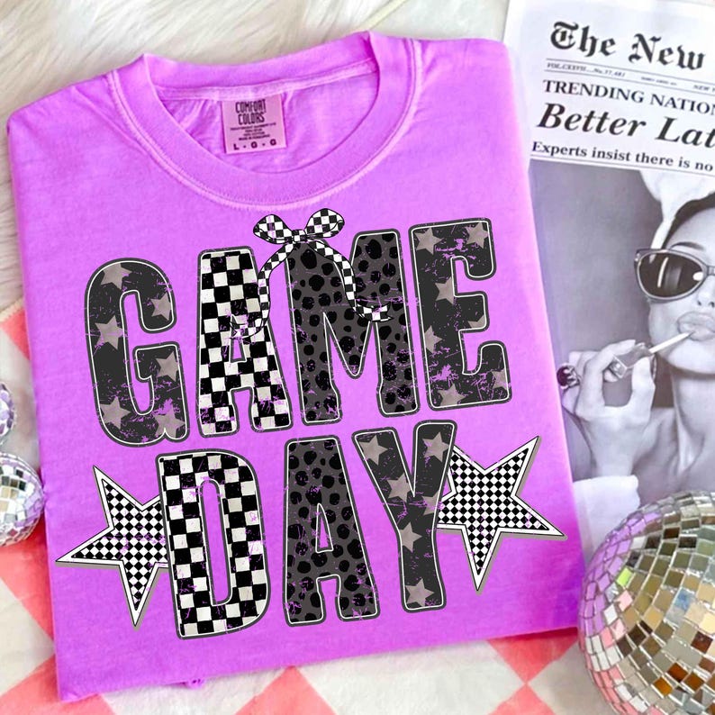 May include: A lilac Comfort Colors t-shirt with the words "GAME DAY" in a mix of black and white checkered and leopard print lettering. The design includes star accents and a checkered bow. A newspaper with the headline "The New" is in the background.