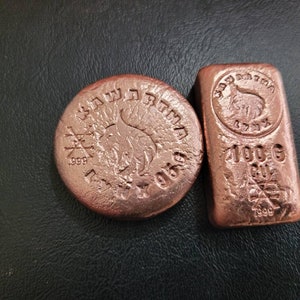 Peut inclure: Deux lingots de cuivre sur une surface sombre. L'un est rond, l'autre rectangulaire. Les deux présentent des motifs et du texte en relief, dont "1999" et "100 G CU". Le lingot rond a un emblème central et du texte sur le bord. Le lingot rectangulaire a également un emblème.