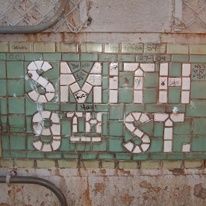 以下が含まれることがあります： 緑と白のタイルを背景に、白いブロック文字で「SMITH 9TH ST.」と書かれた、風化した壁タイルのモザイク。落書きがタイルに見られ、都会的な雰囲気を醸し出しています。