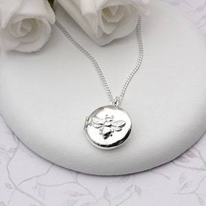 May include: A silver locket necklace featuring a bee design. The locket is round and has a hinged lid. The chain is silver and has a curb link style.
