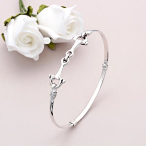 Puede incluir: Un brazalete de plata con un diseño de bocado de caballo. El brazalete está abierto y tiene un cierre con bisagra.