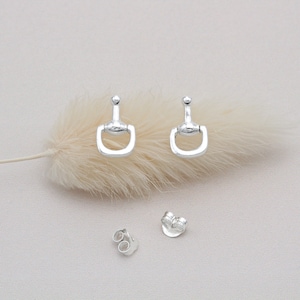 Puede incluir: Un par de pendientes de plata en forma de herradura con una pequeña bola en la parte superior de cada pendiente. Los pendientes están sobre una superficie blanca y esponjosa.