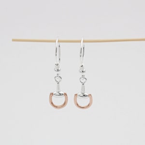 May include: Silver and rose gold horse bit dangle earrings. The earrings are in the shape of a horse bit with a rose gold loop and silver accents.