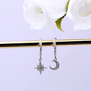 May include: A pair of silver hoop earrings with a dangling star and crescent moon charm. Each charm is encrusted with small, clear crystals.