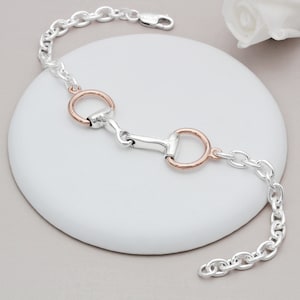 May include: A silver chain bracelet with a rose gold horse bit charm. The bracelet is on a white surface.