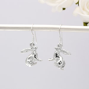 May include: Silver dangle earrings featuring a detailed rabbit design. The rabbit is sitting with its ears up and its tail curled.
