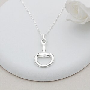 May include: A silver necklace with a snaffle bit pendant. The necklace is on a white surface.