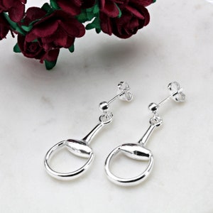 Puede incluir: Un par de pendientes de plata con un diseño de bocado de caballo. Los pendientes tienen una pequeña bola en la parte superior y un abalorio de bocado de caballo redondo más grande que cuelga debajo.