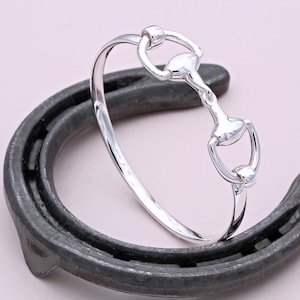 May include: A silver-toned bangle bracelet with a horse bit design. The bracelet is resting on a horseshoe.