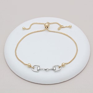 Puede incluir: Un brazalete de cadena dorada con un dije de bocado de caballo plateado. El brazalete es ajustable con un nudo deslizante.