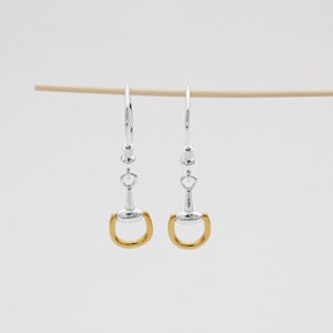 Puede incluir: Un par de pendientes colgantes de plata con dijes de bocado de caballo de color dorado.