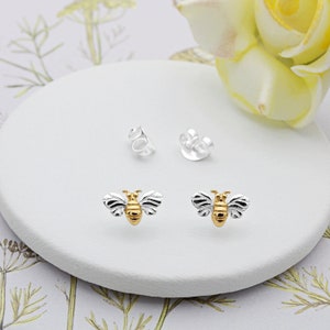 Peut inclure: Une paire de boucles d'oreilles en argent en forme d'abeille avec des accents dorés. Les abeilles sont détaillées avec des ailes et un corps.