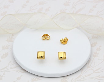 May include: A pair of gold square stud earrings with a smooth, slightly textured surface. The earrings are displayed on a white surface.