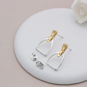 May include: A pair of silver-toned stirrup earrings with gold-toned accents. The earrings are displayed with two pairs of earring backs on a white surface. The design is equestrian-themed.
