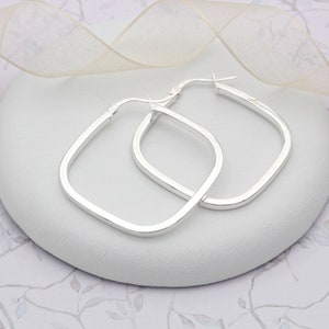 Könnte beinhalten: Ein Paar quadratische Silber-Creolen. Die Ohrringe werden auf einer weißen Oberfläche mit einem weichen, verschwommenen Hintergrund präsentiert. Die Creolen haben ein schlichtes, modernes Design und reflektieren das Licht.