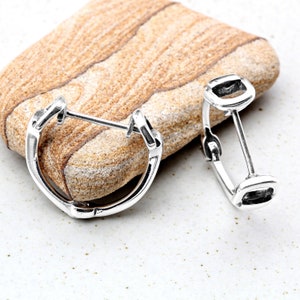 Puede incluir: Un par de pendientes de aro de plata con cierre cuadrado. Los pendientes están sobre una superficie de piedra marrón claro.