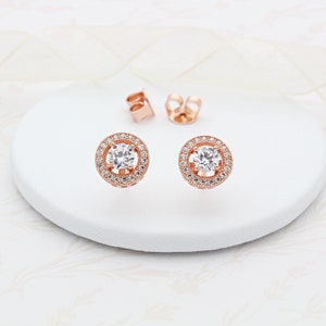 May include: A pair of rose gold stud earrings featuring a round, clear crystal centre surrounded by a halo of smaller crystals.